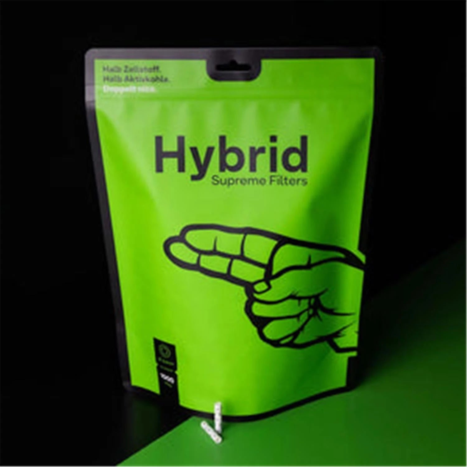 Hybrid Supreme Filters 1000er Standbeutel – leuchtend grüne Großpackung mit schwarzer Hand-Illustration und Aufschrift "Halb Zellstoff. Halb Aktivkohle. Doppelt nice.", vor schwarzem Hintergrund mit grünem Lichtschein, zwei Filter vor der Verpackung sichtbar